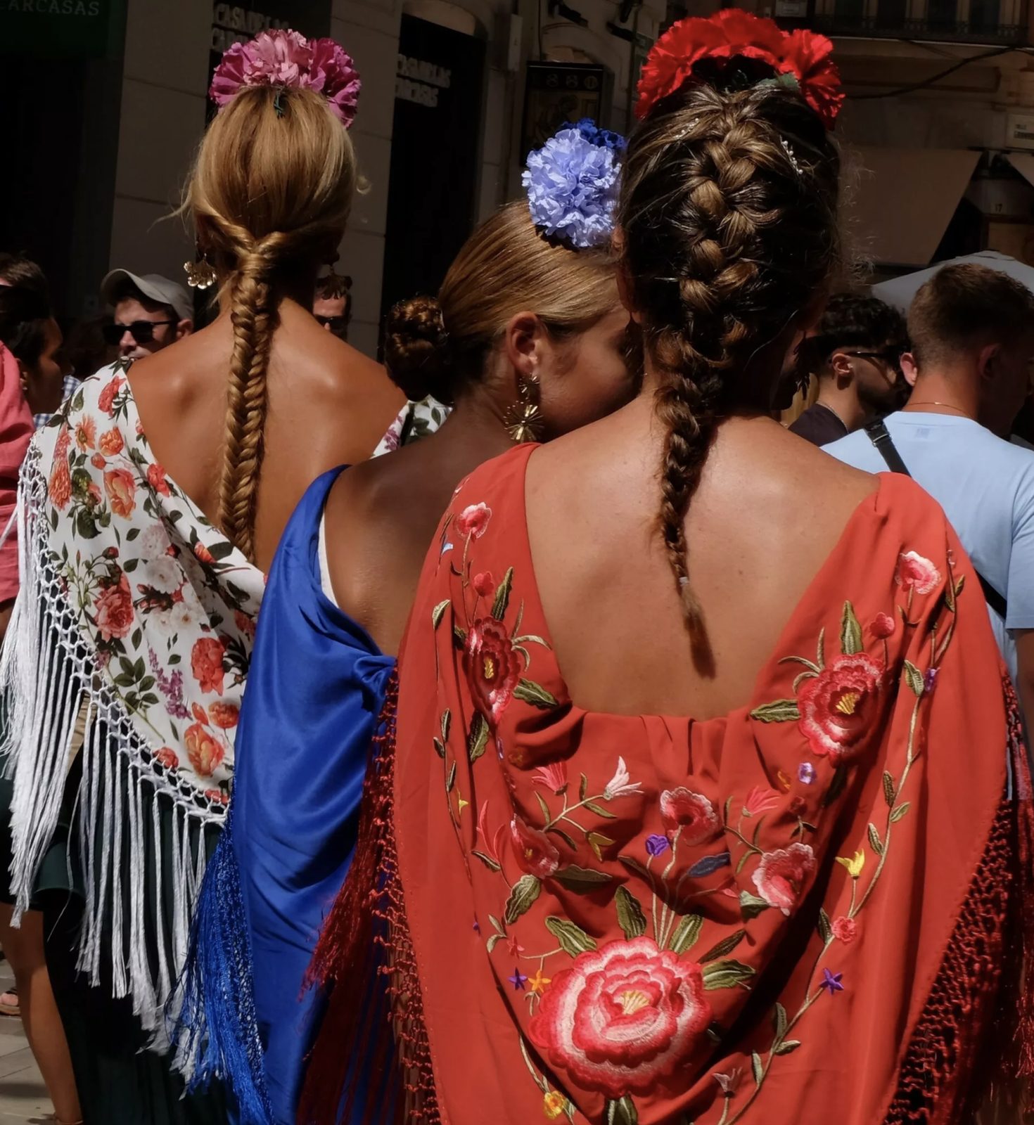 Traditional flamenco dresses with flowers braided into the hair, seen from behind in a Málaga street crowd during Feria